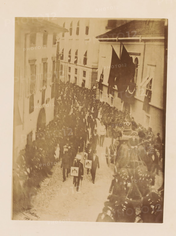 Funeral procession of Henri d'Artois, photograph, page, photographer: A. Vallois, Göritz (Austria), c. 1878 - in or before 1883, A. Vallois, photographer, Steiermark, paper, albumen print, height 208 mm x width 155 mm, cortege, funeral procession, A sepia-toned photograph showing a narrow cobblestone street lined with multi-story buildings and crowded with people. Many individuals are walking in a procession, several wearing dark formal clothing and carrying rectangular framed items held in front of them. On one side a horse-drawn carriage draped in dark fabric and floral arrangements is visible, with figures standing nearby. Dark flags hang from a building above part of the procession. Windows of the buildings overlook the street and people stand along the sidewalks watching. The overall scene is densely packed with figures moving along the street between the buildings.