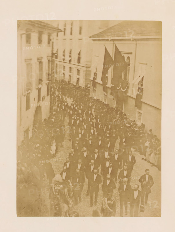 Funeral procession of Henri d'Artois, photograph, page, photographer: A. Vallois, Göritz (Austria), c. 1878 - in or before 1883, A. Vallois, photographer, Steiermark, paper, albumen print, height 211 mm x width 155 mm, cortege, funeral procession, A sepia-toned photograph showing a narrow street crowded with people in formal dark suits and bow ties walking in an organized procession, flanked by onlookers along the sides and leaning from windows of adjacent multi-story buildings, with large dark flags draped from windows on one side and decorative swags hanging beneath some windows, cobblestone pavement underfoot, and ornate uniforms and helmets visible on a few figures near the front of the procession.