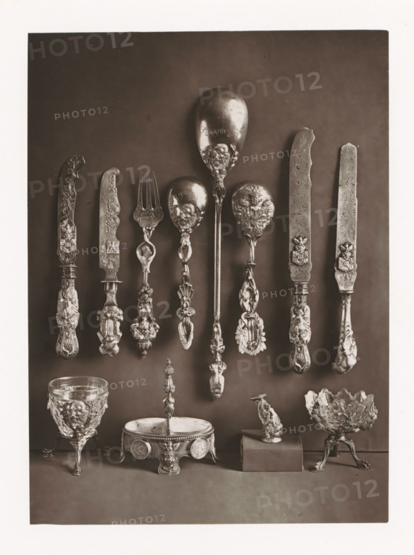Twelve silver objects of the Polish royal house, displayed during the Exposition Rétrospective of 1873 in Tours, France, page, photomechanical print, maker: Gabriel Blaise, Tours, 1873, paper, Woodburytype, Woodburytypie, height 194 mm x width 141 mm, temporary exhibition, 'Salon', table-ware, Period, 1873-05-03 - 1873-05-25, A vertical photograph of ornate metalware arranged against a dark backdrop, showing a row of decorated utensils suspended near the upper half including several knives with elaborately carved handles and blades, a fork with a decorative stem, and multiple spoons with sculpted bowls and ornate handles; below them are decorative objects placed on a surface including a goblet with a raised relief design and tripod feet, a round footed dish with embossed sides and a central finial, a small sculptural figure mounted on a rectangular block, and a bowl with a textured, petal-like edge supported on a decorated tripod base.