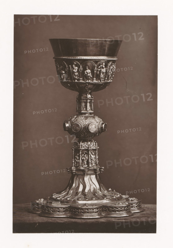 Chalice from a monastery in Tours, installed during the Exposition Rétrospective of 1873 in Tours, France, page, photomechanical print, maker: Gabriel Blaise, Tours, 1873, paper, Woodburytype, Woodburytypepie, height 190 mm x width 125 mm, temporary exhibition, 'Salon', chalice, Period, 1873-05-03 - 1873-05-25, A tall ornate metal chalice is centered against a plain dark background, showing a wide bowl with a smooth inner surface and a decorative band around its lower rim featuring small arched niches each containing a standing human figure in relief; below the bowl a rounded knop with circular medallions and foliate patterns connects to a columnar section decorated with miniature sculpted figures and architectural details, and the base flares into a patterned pedestal with flowing, wave-like ridges and an outer platform adorned with repeating ornamental motifs.