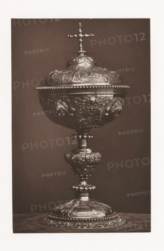 Ciborium of precious metal, installed during the Exposition Rétrospective of 1873 in Tours, France, page, photomechanical print, maker: Gabriel Blaise, Tours, 1873, paper, Woodburytype, Woodburytypepie, height 196 mm x width 126 mm, temporary exhibition, 'Salon', ciborium, Period, 1873-05-03 - 1873-05-25, A tall ornate chalice with a hinged domed lid topped by a small cross sits against a plain dark background, the body and stem covered in intricate raised floral and scrollwork decorations and small sculpted faces, the base broad and heavily ornamented with layered rosettes and beaded edges, the metal surface showing highlights and shadows that emphasize the embossed details.
