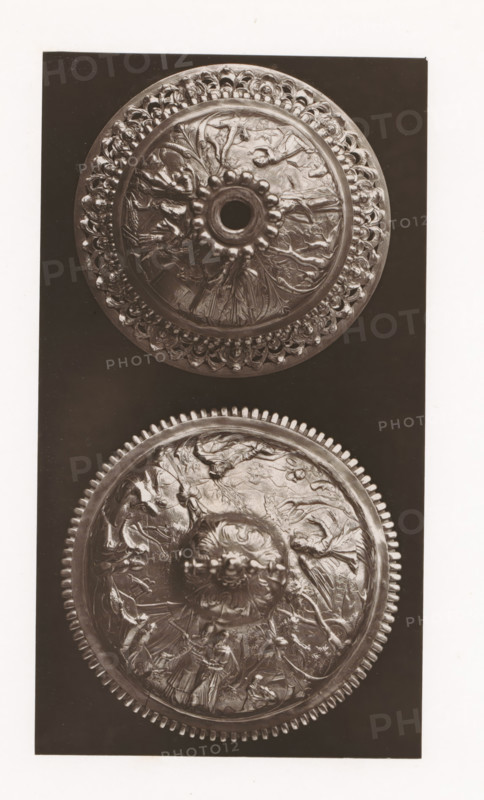 Two ciborium lids, installed during the 1873 Exposition Rétrospective in Tours, France, page, photomechanical print, maker: Gabriel Blaise, Tours, 1873, paper, Woodburytype, Woodburytypepie, height 202 mm x width 111 mm, temporary exhibition, 'Salon', ciborium, Period, 1873-05-03 - 1873-05-25, A vertical photograph shows two round, ornate metal lids or medallions set against a dark rectangular background and surrounded by a light border. Each round piece features a raised central knob with petal-like protrusions around a central opening on the upper one and a carved floral boss on the lower one. Both discs display intricate relief scenes with human figures, animals, drapery, and flowing organic motifs radiating outward, and each rim is embellished with repeating decorative elements; the upper rim has a scalloped, bead-like pattern while the lower rim shows a toothed, gear-like border. The metal surfaces exhibit highlights and shadows that emphasize the embossed details and textured wor...
