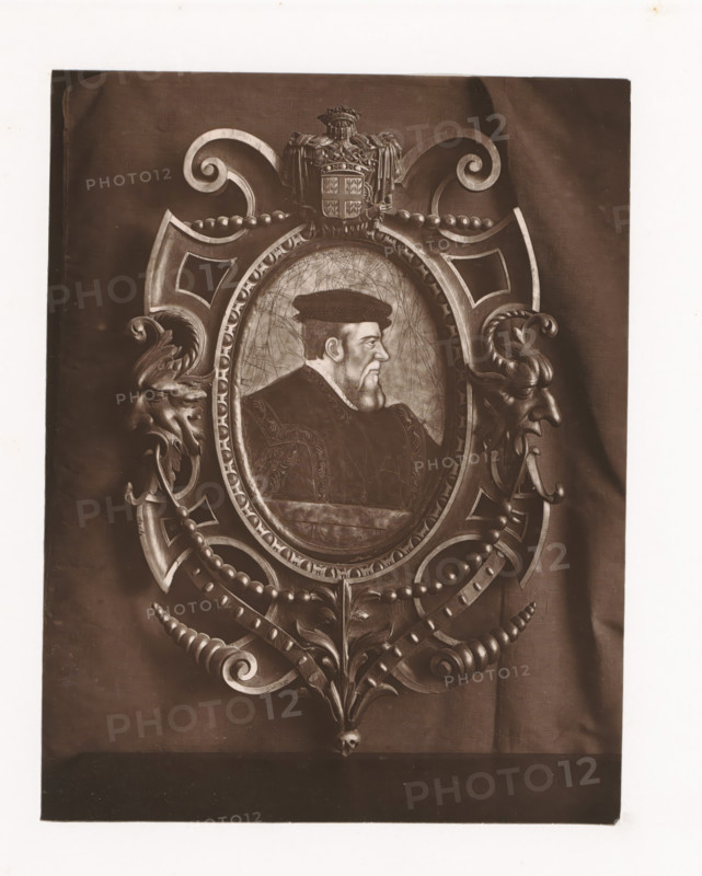 Portrait of Anne de Montmorency in a decorated frame, mounted during the Exposition Rétrospective of 1873 in Tours, France, page, photomechanical print, maker: Gabriel Blaise, Tours, 1873, paper, Woodburytype, Woodburytypepie, height 170 mm x width 131 mm, Person, Anne de Montmorency, historical persons, picture, painting, temporary exhibition, 'Salon', Period, 1873-05-03 - 1873-05-25, A framed oval portrait of a bearded man in profile wearing a cap and ornate clothing sits at the center of an elaborate carved wooden surround. The surround features scrolling volutes, beaded garlands, and symmetrical decorative elements including stylized faces with curled horns on either side. Above the oval is a crest with draped fabric and ornamental detailing, and below the frame are acanthus-like leaves and curved, ribbed scrolls ending in a small skull motif. The image has a sepia tone and the background shows a dark draped cloth behind the framed composition.