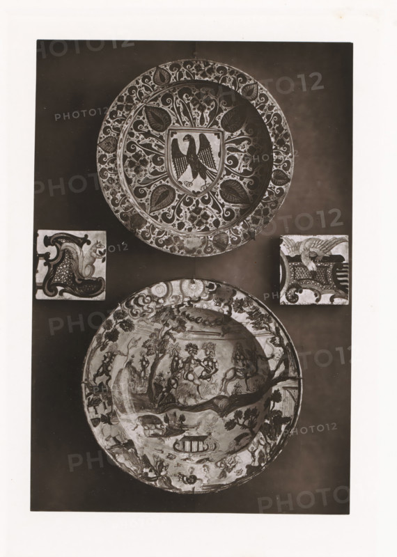Two bowls and two earthenware tiles, displayed during the Exposition Rétrospective of 1873 in Tours, France, page, photomechanical print, maker: Gabriel Blaise, Tours, 1873, paper, Woodburytype, Woodburytypepie, height 194 mm x width 132 mm, temporary exhibition, 'Salon', dish, plate, saucer, Period, 1873-05-03 - 1873-05-25, A vertical photograph shows two large decorative plates and two square tiles arranged on a dark background, the upper plate featuring an ornate floral border and a central shield with a stylized bird, the lower plate displaying a busy scene with many small figures and landscapes around its rim, and the two square tiles positioned between the plates each containing different ornamental motifs including a curled decorative shape on one and a winged creature on the other.