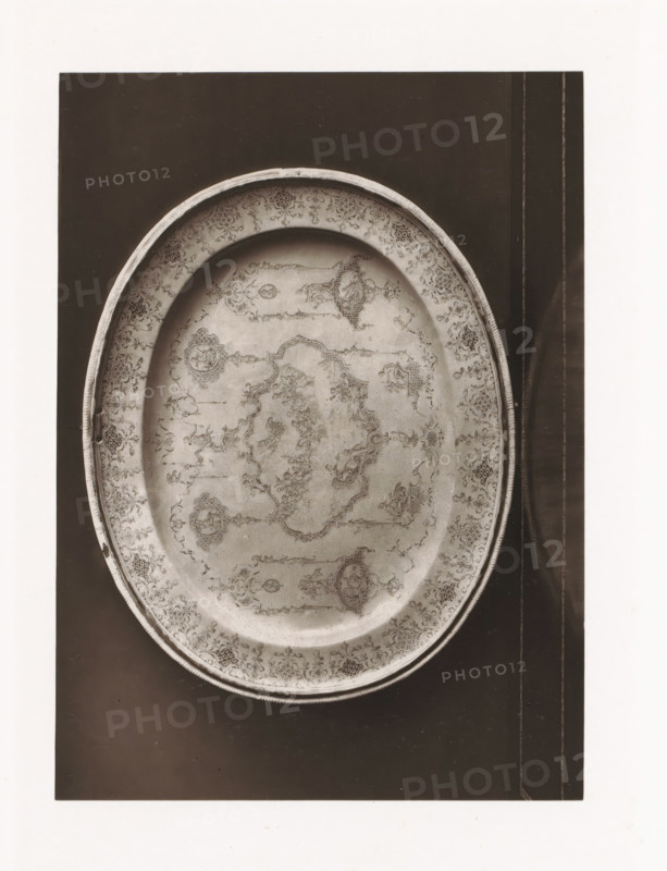 Small earthenware bowl from a monastery, displayed during the Exposition Rétrospective of 1873 in Tours, France, page, photomechanical print, maker: Gabriel Blaise, Tours, 1873, paper, Woodburytype, Woodburytypepie, height 165 mm x width 120 mm, temporary exhibition, 'Salon', dish, plate, saucer, Period, 1873-05-03 - 1873-05-25, An oval metal tray with an ornate etched design fills most of the image, showing a raised rim with a patterned border and an interior decorated with intricate floral and geometric motifs and symmetrical medallions; the tray rests on a dark rectangular surface with narrow lighter vertical lines near the right edge, and a white margin surrounds the dark background.