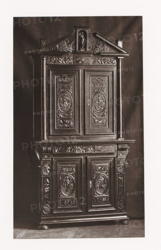 Cabinet from Château d'Anet, installed during the Exposition Rétrospective of 1873 in Tours, France, page, photomechanical print, maker: Gabriel Blaise, Tours, 1873, Gabriel Blaise, paper, Woodburytype, Woodburytypepie, height 197 mm x width 116 mm, temporary exhibition, 'Salon', ornamental cupboard (wood and other phytogenic material), Period, 1873-05-03 - 1873-05-25, A tall dark wooden cabinet with ornate carved panels and decorative pilasters, featuring upper and lower sections with framed doors, oval relief medallions depicting human figures, floral and scroll motifs, a triangular pediment at the top with a central niche holding a carved standing figure, carved horizontal friezes with reclining figures, fluted columns on either side, molded base and rounded feet, and a dark backdrop behind the cabinet.