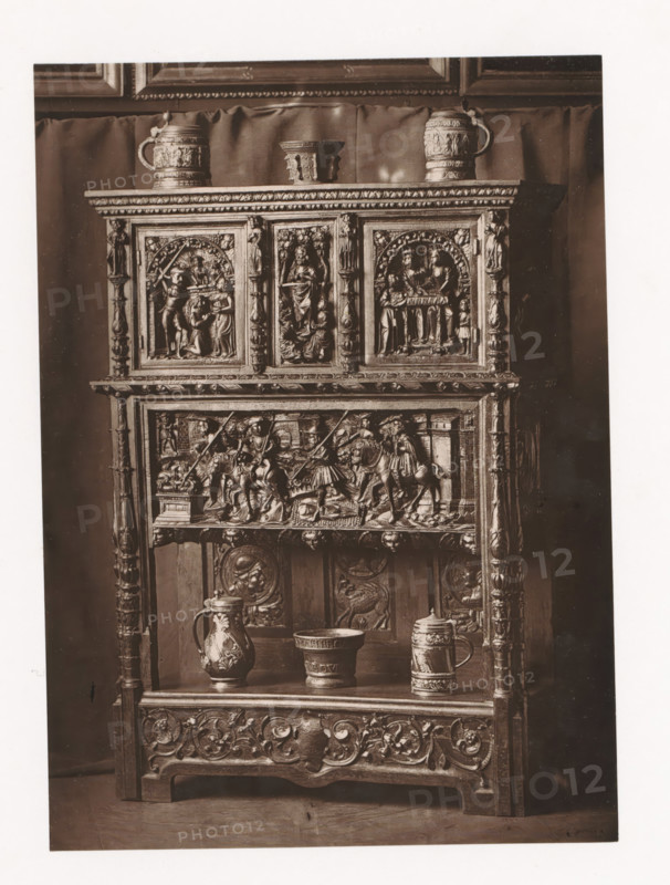 Sideboard in the style of Francis I of France, displayed during the Exposition Rétrospective of 1873 in Tours, France, page, photomechanical print, maker: Gabriel Blaise, Tours, 1873, paper, Woodburytype, Woodburytypie, height 179 mm x width 130 mm, temporary exhibition, 'Salon', ornamental cupboard (wood and other phytogenic material), Period, 1873-05-03 - 1873-05-25, A tall wooden cabinet with ornate carved panels and columns fills the frame, its upper section featuring three framed relief scenes set beneath a carved cornice, with a small pitcher and a lidded tankard placed on top and a small cup between them, the middle register showing a detailed horizontal relief of figures and horses within an architectural setting, and the lower shelving holding a decorated jug, a wide bowl, and a tall tankard, all set against a draped backdrop and photographed in monochrome.