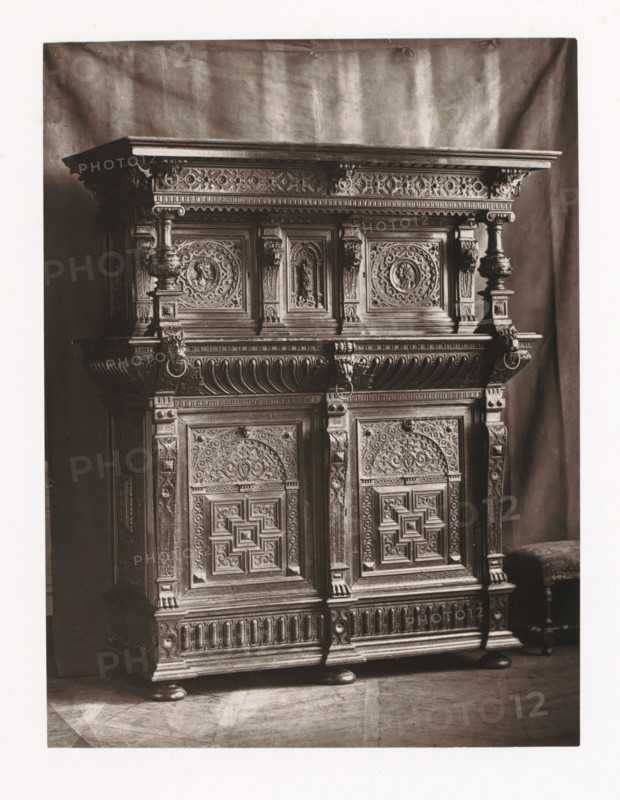 Flemish style sideboard, installed during the 1873 Exposition Rétrospective in Tours, France, page, photomechanical print, maker: Gabriel Blaise, Tours, 1873, paper, Woodburytype, Woodburytypepie, height 213 mm x width 161 mm, temporary exhibition, 'Salon', ornamental cupboard (wood and other phytogenic material), Period, 1873-05-03 - 1873-05-25, A large ornate wooden cabinet fills the frame, showing the upper section and doors with elaborate carved panels and decorative molding; the top has a flat projecting cornice with carved frieze beneath, supported by carved columns and brackets featuring rounded baluster shapes and carved lion head rings, while the door panels display intricate geometric and floral reliefs surrounded by carved borders and small columnar pilasters; the lower section shows carved vertical fluting and rounded feet resting on a patterned floor, and a small upholstered stool with turned legs is visible at the right edge against a draped backdrop.