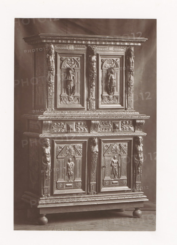 Cabinet in the style of Henry III of France, installed during the Exposition Rétrospective of 1873 in Tours, France, page, photomechanical print, maker: Gabriel Blaise, Tours, 1873, paper, Woodburytype, Woodburytypepie, height 184 mm x width 123 mm, temporary exhibition, 'Salon', ornamental cupboard (wood and other phytogenic material), Period, 1873-05-03 - 1873-05-25, The image shows a tall wooden cabinet with ornate carved panels and decorative molding, featuring sculpted human figures in recessed rectangular frames, fluted pilasters with carved faces and foliage, a projecting cornice with carved geometric and leaf patterns, a middle shelf with scrollwork and raised relief motifs, paneled doors with central standing figures, carved rosettes and acanthus leaves, and the cabinet resting on rounded bun feet on a wooden floor.