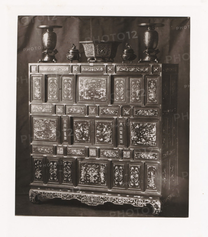 Cabinet from Annam, installed during the Exposition Rétrospective of 1873 in Tours, France, page, photomechanical print, maker: Gabriel Blaise, Tours, 1873, paper, Woodburytype, Woodburytypepie, height 178 mm x width 154 mm, temporary exhibition, 'Salon', ornamental cupboard (wood and other phytogenic material), Period, 1873-05-03 - 1873-05-25, A tall wooden cabinet fills the frame, its surface covered in numerous rectangular panels each decorated with intricate painted scenes and floral motifs; the cabinet rests on a carved base with geometric fretwork and curved feet. On top of the cabinet sit several metal vessels including two tall urns with wide flat lids positioned near the outer edges, a large square-shaped bowl with ornate relief work in the center, and two smaller lidded containers with sculpted figures on their lids flanking the central bowl. The background is a plain dark cloth that contrasts with the lighter tones of the cabinet and metal objects.