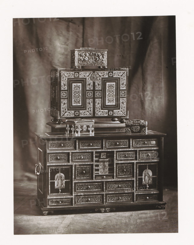 Two cabinets and three chests, displayed during the 1873 Exposition Rétrospective in Tours, France, page, photomechanical print, maker: Gabriel Blaise, Tours, 1873, paper, Woodburytype, Woodburytypepie, height 206 mm x width 156 mm, temporary exhibition, 'Salon', chest, coffer (wood and other phytogenic material), ornamental cupboard (wood and other phytogenic material), Period, 1873-05-03 - 1873-05-25, A photograph showing a multi-tiered ornate cabinet with intricate inlaid patterns and decorative panels placed against a draped fabric backdrop, the cabinet featuring a larger lower section with many small drawers and framed panels, a central open compartment with a carved figure, ornate metal ring handles on the sides, and raised feet, while a smaller decorated box sits atop the cabinet and an additional rectangular box with carved floral motifs rests above that, all finished with detailed filigree and contrasting light and dark surfaces.