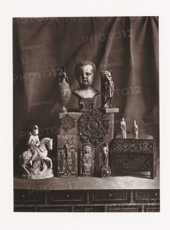 Various objects, displayed during the Exposition Rétrospective of 1873 in Tours, France, page, photomechanical print, maker: Gabriel Blaise, Tours, 1873, paper, Woodburytype, Woodburytypepie, height 165 mm x width 124 mm, temporary exhibition, 'Salon', Period, 1873-05-03 - 1873-05-25, A display of sculptural and carved objects arranged on a surface in front of a draped fabric backdrop includes a central bust of a child's head on a pedestal, flanked by an ornate vase with a narrow neck and a tall fragmented figure on a base, a round decorative medallion with elaborate foliage detailing mounted on a box, two small standing figurines on a raised box to the right, and a carved chest with geometric and floral patterns; below and in front are a mounted knight on horseback poised above a prone figure, several vertical carved panels depicting seated and standing figures and ornamental motifs, and a small column-like figure on a square base, all positioned on top of a richly decorated cabinet w...