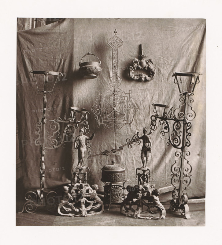 Various objects, displayed during the Exposition Rétrospective of 1873 in Tours, France, page, photomechanical print, maker: Gabriel Blaise, Tours, 1873, paper, Woodburytype, Woodburytypepie, height 181 mm x width 165 mm, temporary exhibition, 'Salon', Period, 1873-05-03 - 1873-05-25, A sepia-toned photograph shows a backdrop of draped fabric with decorative metal objects arranged in front of it, including ornate wrought iron stands with scrollwork on either side, a hanging bowl with embossed patterns suspended by a handle, a tall decorative metal panel with intricate openwork and a pointed finial, and a carved ornamental wreath with sculpted animal heads. In the lower area there are sculpted figurines mounted on bases, a lidded cylindrical container with embossed bands, multiple intricately carved supports featuring seated cherubic figures, and connecting metal rods and chains linking some of the elements together.