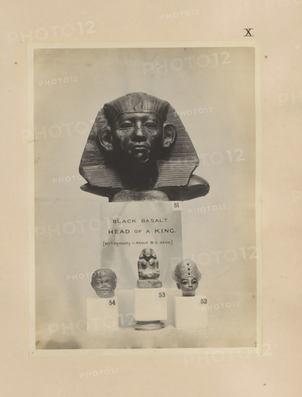 Four Egyptian objects, including a fragment of a sculpture, the head of a pharaoh and a sculpture of a seated scribe, photograph, page, number: 'XII', c. 1890 - c. 1895, photographer, paper, gelatin silver print, developed gelatin silver print, height 213 mm x width 157 mm, sculpture, king, writer, poet, author, A photograph shows a dark sculpted head wearing a striped headpiece with flared sides and a smooth face with prominent ears, positioned above three smaller sculpted heads on pedestals; the central smaller sculpture is a seated figure on a stepped base, flanked by a rough-textured head on the left and a head with a patterned headdress on the right, all set against a pale background and framed by a lighter rectangular border within a larger mottled sheet.