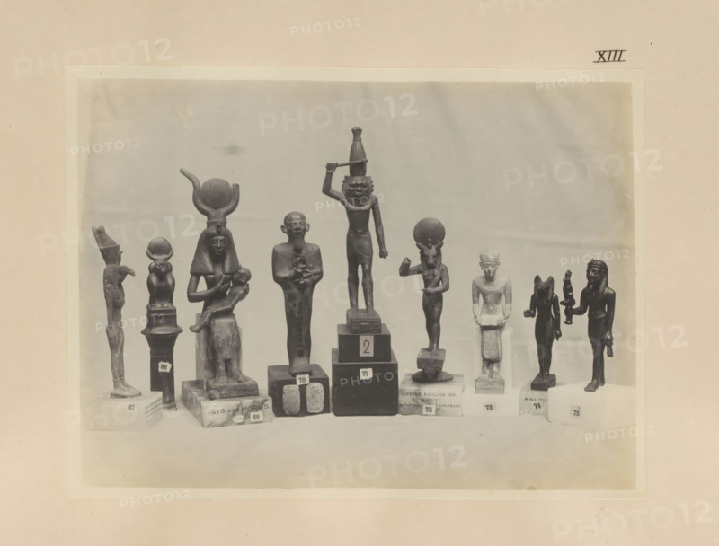 Nine Egyptian objects, including bronze sculptures of Mut, Anupt, Isis with Horis and Apis, photograph, page, number: 'XIII', c. 1890 - c. 1895, photographer, paper, gelatin silver print, developing gelatin silver print, height 158 mm x width 213 mm, sculpture, representations, Egyptian gods, demi-gods, heroes, etc. (with NAME), Horus, son of Osiris and Isis, Isis, the mother-goddess, Egyptian goddesses, demi-goddesses, heroines, A row of small dark statues and figurines stands on a flat surface against a light background, each mounted on individual pedestals of varying shapes and heights; the figures include humanlike forms with headdresses and seated postures, a figure holding a smaller figure, a standing figure with an arm raised and another with a circular element above its head, a taller central figure with an arm raised holding a horizontal object and a carved face, and several smaller standing figures with distinct headgear and limbs posed differently, all appearing in a uniform...