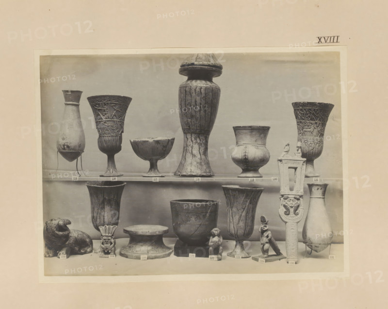 Sixteen Egyptian objects, vases, a sculpture of Bes and a sistrum, photograph, page, number: 'XVIII', c. 1890 - c. 1895, photographer, paper, gelatin silver print, developing gelatin silver print, height 158 mm x width 213 mm, sculpture, containers, representations, Egyptian gods, demi-gods, heroes, etc. (BES), A black and white photograph of a display of various ancient-style vessels and small sculptures arranged on shelves, including tall fluted goblets with carved figures, bulbous jars with narrow necks, shallow bowls on stems, a large tall vase with a rounded lid, a carved column-like object with decorative figures, a seated animal sculpture, small human or animal figurines, and several pedestal bases, all shown against a plain background.