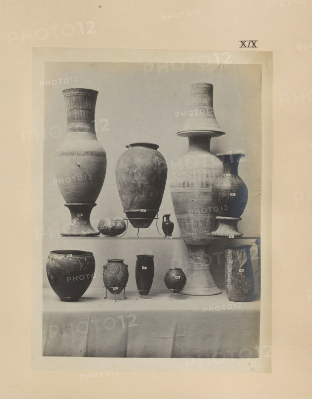 Eleven Egyptian vases, photograph, page, number: 'XIX', c. 1890 - c. 1895, photographer, paper, gelatin silver print, developing gelatin silver print, height 208 mm x width 155 mm, containers, A centered photograph mounted on a pale sheet shows a display of ceramic vessels arranged on shelves draped with fabric; the top shelf holds a variety of tall urns and amphorae with rounded bodies, flared rims, narrow necks, and pedestal bases in muted tones and worn surfaces, including a large bulbous vessel, a tall slender vase with a wide flaring neck, and a conical-necked jar, while the lower shelf shows smaller pots and jars of different shapes including globular and cylindrical forms, one with spiral decorative motifs and another with vertical leaf-like patterns, several supported on small metal stands; the overall image has a soft, slightly faded monochrome appearance and visible texture and wear on the ceramics and paper.