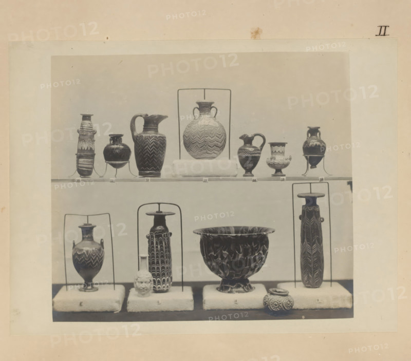 Eleven Egyptian vases and a cup and a pot, photograph, page, number: 'II', c. 1890 - c. 1895, photographer, paper, gelatin silver print, developed gelatin silver print, height 154 mm x width 205 mm, sculpture, container of glass: bottle, jar, vase, container of stone: stone vase, A framed photograph shows a display of various pottery pieces arranged on padded stands and metal supports against a plain backdrop; the visible vessels include tall slender amphorae with patterned surfaces, handled jugs with spouts and zigzag or wave-like designs, a large bowl with a mottled or marbled interior, small rounded jars with ornate surface decoration, and a sculpted head object placed on one of the stands, all in muted tones and photographed in a museum-style arrangement.