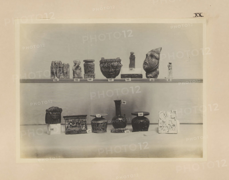 Fourteen Egyptian objects, including a sculpture of Harpocrates and four cabbage pots, photograph, page, number: 'XX', c. 1890 - c. 1895, photographer, paper, gelatin silver print, developing gelatin silver print, height 160 mm x width 212 mm, sculpture, containers, representations, Egyptian gods, demi-gods, heroes, etc. (HARPOCRATES), A shelf display of various small sculptural objects and vessels arranged in two rows against a plain background, including small carved figures, a fragmented human-like head, cylindrical and rounded pots with decorative relief, a carved rectangular box, a lion-like carved block, a tall narrow-necked vase, and a rectangular carved slab, all shown in monochrome with subtle shadows on the shelf and base.