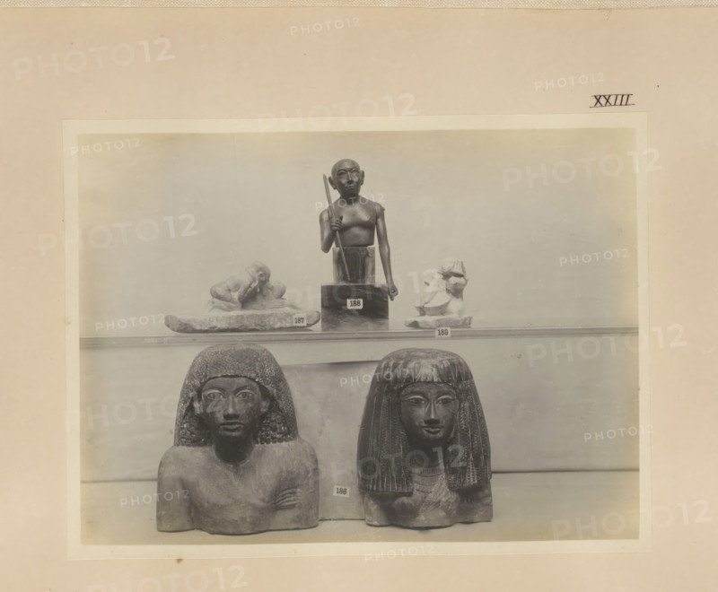 Five Egyptian objects, including two busts, photograph, page, number: 'XXIII', c. 1890 - c. 1895, photographer, paper, gelatin silver print, developing gelatin silver print, height 159 mm x width 214 mm, sculpture, adult woman, adult man, A faded photograph mounted on a beige background shows a display of carved stone figures on a shelf, with a smaller standing figure holding a staff placed above two seated busts below; the seated busts have textured hair or wigs and detailed facial features, while small fragmentary sculptures flank the standing figure on the upper shelf.