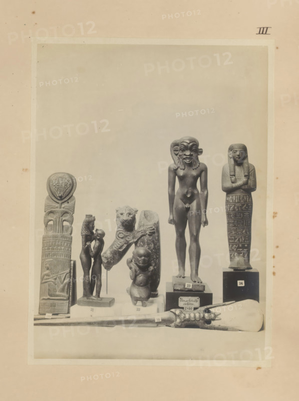 Seven Egyptian objects, including a bronze sculpture of Isis with Horus, a bronze sculpture of Harpocrates with the head of Bes and a wooden ushabti, photograph, page, number: 'III', c. 1890 - c. 1895, photographer, paper, gelatin silver print, developed gelatin silver print, height 212 mm x width 157 mm, sculpture, Horus, son of Osiris and Isis, Isis, the mother-goddess, representations, Egyptian gods, demi-gods, heroes, etc. (BES), A sepia-toned photograph shows a display of small statues and carved objects arranged on pedestals and a flat surface. From left to right there is a tall vertical carved pillar topped with a round emblem, a slender figurine of a standing pair with one figure slightly smaller and facing the taller figure, a sculpted animal head mounted on a fragment of carved stone beside a small rounded figure with a childlike form, a taller nude male figure with an exaggerated facial mask standing on a square base, and a mummiform figure with arms crossed and detailed pat...