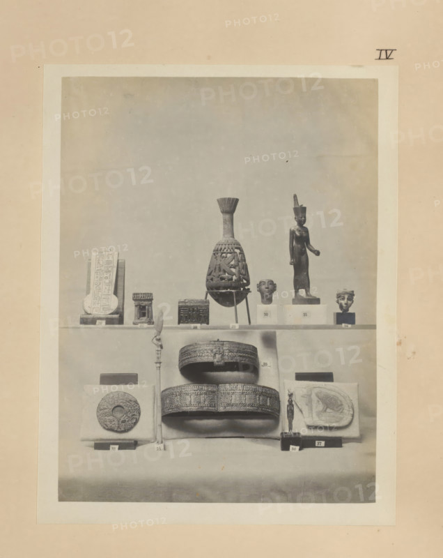 Twelve Egyptian objects, including a bronze sculpture of Neith, two gold bracelets and a gold sculpture of Hathor, photograph, page, number: 'IV', c. 1890 - c. 1895, photographer, paper, gelatin silver print, developing gelatin silver print, height 203 mm x width 158 mm, sculpture, bracelet, wristlet, representations, Egyptian goddesses, demi-goddesses, heroines, etc. (NEITH), A framed black-and-white photograph of a display shelf filled with small artifacts, centered on a tall decorative vessel with a narrow neck and flared top, flanked by a standing figurine wearing a tall headdress and a sculpted head on simple rectangular plinths, with a carved rectangular tablet and several small box-like objects on the left side, and a small sculpted head on a plinth on the right; below the shelf are two ornate circular boxes stacked above a rounded base, a tall slender staff with a figure near its top, a round carved disk mounted on a cushion, a small standing figurine on a square base, and a re...