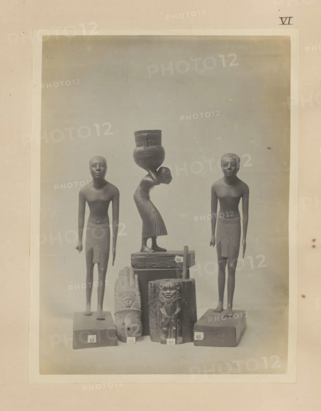 Five Egyptian objects, including two sculptures of boys and a wooden kohl pot with the image of Bes, photograph, page, number: 'VI', c. 1890 - c. 1895, photographer, paper, gelatin silver print, developed gelatin silver print, height 212 mm x width 161 mm, sculpture, youth, adolescent, wooden container: barrel, cask, bucket, representations, Egyptian gods, demi-gods, heroes, etc. (BES), A framed photograph shows a group of carved figures arranged on a flat surface against a neutral backdrop, including two standing male figures with short hair and wrapped skirts positioned on either side, a central female figure bent forward carrying a large decorated vessel on her head, several smaller carved objects and blocks with relief faces and patterns stacked in front, and a pale border surrounding the image with a small mark near the top right.