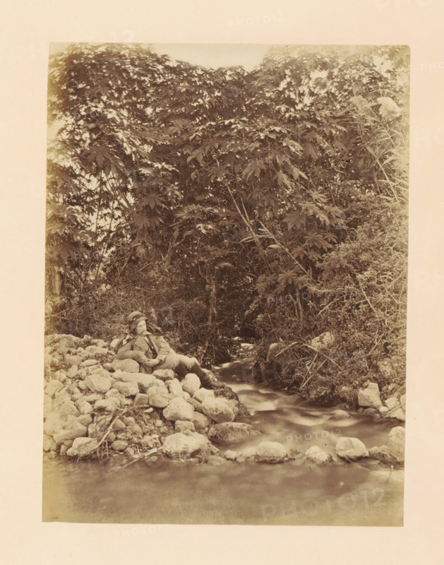 View of Elisha's Fountain in Jericho, photograph, page, photographer: Horatio Herbert Kitchener, Jericho, c. 1870 - c. 1875, Horatio Herbert Kitchener, photographer, Palestine, West Bank, paper, albumen print, height 200 mm x width 151 mm, spring, source of river or stream, A sepia-toned photograph showing a rocky stream flowing from the middle toward the bottom foreground, with smooth water over and between rounded stones; dense foliage and tall leafy plants rise behind the stream, creating a dark textured backdrop of leaves and branches; a person wearing a hat and coat reclines on a pile of rocks at the stream edge, leaning on one elbow and looking slightly upward, with legs extended and one knee bent; shrubs and grasses grow along the stream banks and some branches and sticks lie among the stones.