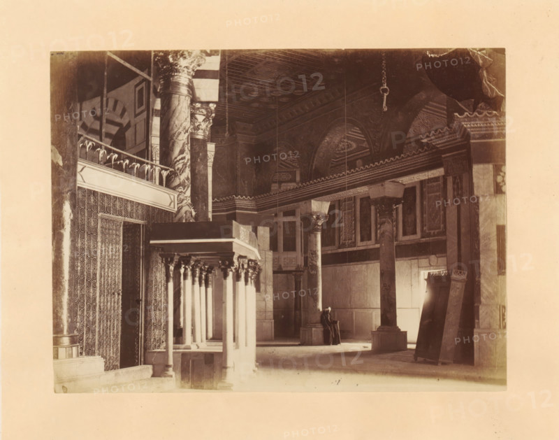 Interior of the Dome of the Rock on the Temple Mount, photograph, page, photographer: Horatio Herbert Kitchener, Dome of the Rock, c. 1870 - c. 1875, Jerusalem, paper, albumen print, height 155 mm x width 206 mm, temple, shrine, Islam, Mohammedanism, A sepia photograph of an interior space showing tall decorative columns with ornate capitals and marble-like surfaces, arched ceilings with patterned coffers, and a row of slender columns supporting a balcony or canopy with carved details; patterned wall coverings and carved panels line one side, a large rectangular column or pulpit structure stands in the foreground, and a person seated on a bench wearing dark clothing and a head covering is visible near the back beneath the arches.