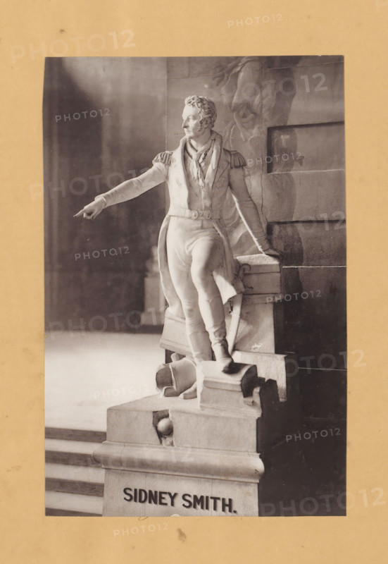 Shooting of the statue of Sidney Smith, page, photomechanical print, c. 1876 - in or before 1881, paper, collotype, photoprint, height 211 mm x width 139 mm, Person, Sidney Smith, historical persons, sculpture, A monochrome photograph of a marble statue mounted on a stone pedestal showing a standing male figure in a historical military uniform with epaulets and a sash, one arm extended outward with a pointed finger and the other resting on a block, the figure wearing a long coat and fitted trousers with boots, curly hair and a turned profile, carved drapery and architectural stone blocks behind the statue, visible steps leading up beside the pedestal, and a plain border around the photograph.