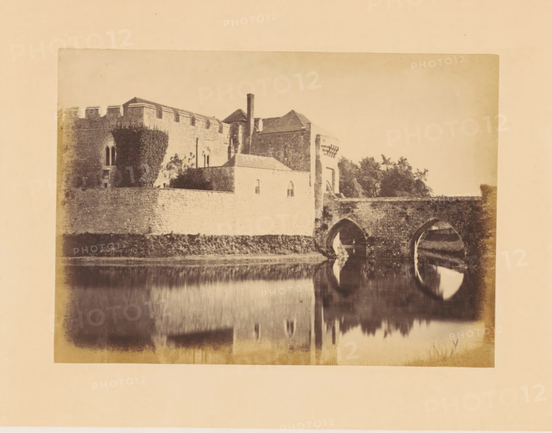Gatehouse and bridge of Leeds Castle, photograph, page, inscription: 'No.4.', photographer: John Cruttenden, Leeds Castle, c. 1864 - in or before 1869, Leeds, paper, albumen print, height 144 mm x width 201 mm, landscape with tower or castle, castle, gate, entrance, A sepia-toned photograph of a stone building complex with crenellated walls and arched windows beside a bridge with two large stone arches spanning a calm body of water that reflects the arches and parts of the structure, with ivy growing on a section of the wall and trees visible behind the building.