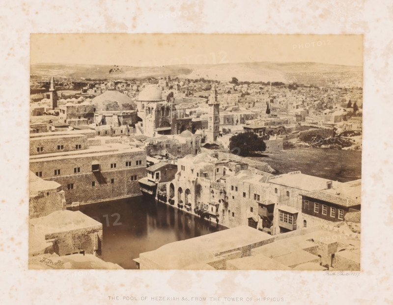 View of Jerusalem, The pool of Hezekiah &c_ from the tower of Hippicus, page, This photo is part of a cover with 7 sheets of albumen prints., inscription: 'Jerusalem', photographer: Francis Frith, Jerusalem, 1857, paper, albumen print, height 161 mm x width 228 mm, Person, city-view, and landscape with man-made constructions, A sepia-toned panoramic view of an old city with stone buildings and domed roofs clustered across the landscape, a body of dark reflective water bordered by stone walls and arches in the foreground, narrow balconies and small windows dotting the facades above the water, a tall slender tower rising among the buildings, scattered trees and a larger dark tree near the water's edge, rolling hills visible in the distance, and a mottled paper border surrounding the image.