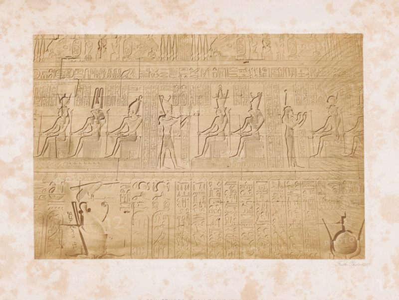 Hieroglyphics on the outer wall of the temple complex at Denderea, Sculptures from the outer wall, page, This photo is part of a cover with 7 pages of albumen prints., inscription: 'Dendera', photographer: Francis Frith, Dendera, 1857, North Africa, paper, albumen print, height 151 mm x width 223 mm, Person, temple, shrine, Egyptian religion, Egyptian hieroglyphs, Dendera Temple complex, A rectangular stone panel with carved figures and decorative bands sits against a mottled light background; the panel shows seated humanlike figures in profile wearing tall headdresses and holding staffs, alternating with standing figures in profile raising arms or holding objects, all rendered in shallow bas relief with detailed headdresses, garments, and stylized limbs; the upper portion contains horizontal registers filled with repeated narrow vertical motifs and small carved animals and symbols, while the lower area displays more compact vertical columns of carved motifs and scenes including a larg...