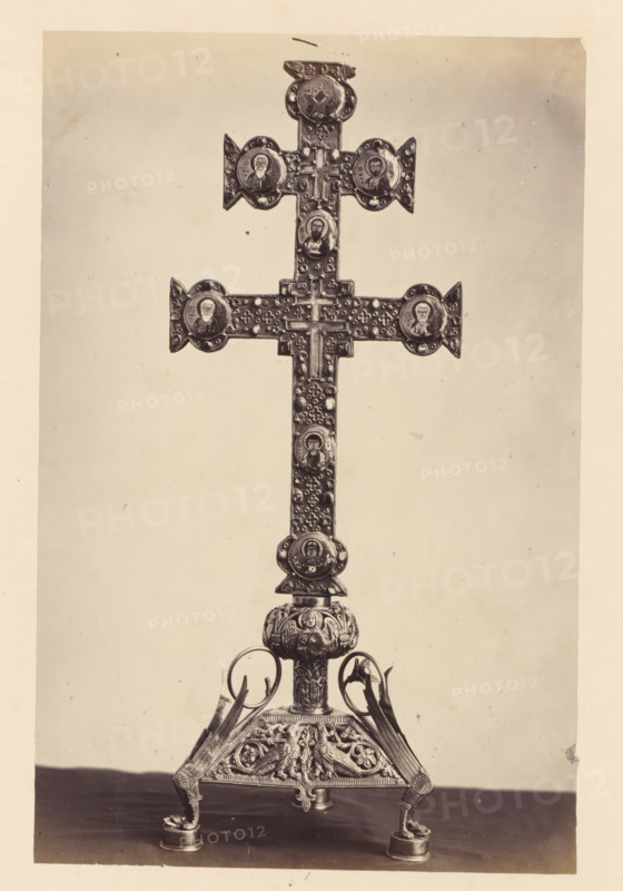 Golden cross with relic from the collection of the Soeurs de Notre-Dame de Namur, displayed at an exhibition of religious objects from the Middle Ages and Renaissance in 1864 in Mechelen, photograph, page, number: '1', photographer: Joseph Maes, Mechelen, 1864, paper, albumen print, height 269 mm x width 175 mm, relics (and reliquaries), piece of sculpture (precious metals, arts), Period, A tall ornate metal cross mounted on an elaborately decorated stand fills the frame, with a vertical shaft and a transverse beam forming a cross shape adorned with circular medallions featuring portrait-like images and textured decorative elements; the vertical shaft shows repeated small raised motifs and additional medallions, and the top and ends of the horizontal arm have pointed triangular extensions. The stand beneath the cross is highly detailed with sculpted figures and winged motifs, curved supports rising to meet the central column, and small rounded feet resting on a flat surface. The backgr...