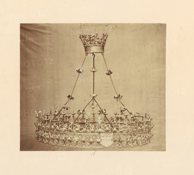 Two metal candlesticks with bases, displayed at an exhibition of religious objects from the Middle Ages and Renaissance in 1864 in Mechelen, photograph, page, number: '17 - 18', photographer: Joseph Maes, Mechelen, 1864, paper, albumen print, height 258 mm x width 196 mm, chandelier, candelabrum, Period, A large ornate metal crown suspended by slender rods is visible against a plain backdrop, the crown featuring a circular base with repeated floral and arched decorative motifs, numerous small cup-like holders around the rim, vertical supports rising to a narrower crown-like top with intricate openwork and fleur-de-lis shaped elements, and decorative rings and curved connectors along the supports; the image has a wide margin of blank paper surrounding the photographed crown.