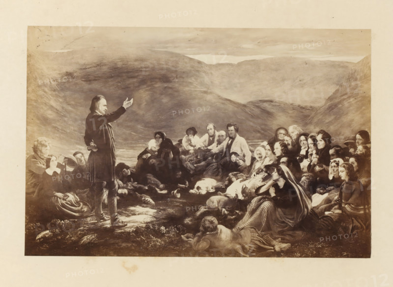 Photo reproduction of Sabbath in the Glen by Sir George Harvey, photograph, reproduction, page, photographer: Thomas Annan, after painting by George Harvey (Sir), c. 1865 - in or before c. 1870, paper, albumen print, height 146 mm x width 220 mm, meeting, assembly - AA - in the open air, Sabbath, Jewish religion, Scotland, A wide sepia-toned scene shows a standing man in dark clothing with one arm raised and a small book at his side facing a large seated and reclining crowd; the crowd includes men, women, and children in period-style clothing with shawls, bonnets, and coats, several people looking toward the standing man while others look outward or downward, and a few individuals appear to be resting or asleep; the foreground features rocks, grass, and a reclining figure facing away from the viewer, and a few animals lying on the ground; the background displays rolling hills and a cloudy sky.