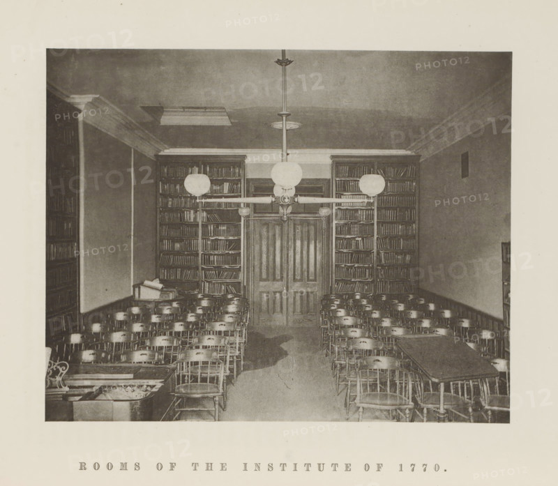 Rooms of the Institute of 1770, Interior of the Institute of 1770 of Harvard University, page, photomechanical print, c. 1870 - in or before 1875, paper, collotype, photoprint, height 164 mm x width 205 mm, university building, college, club (students), North America, Massachusetts, Cambridge (MA), Harvard College, A sepia-toned interior photograph showing a room with rows of wooden chairs arranged facing a central double door flanked by tall bookshelves filled with books, overhead light fixtures with round globe shades suspended from the ceiling, a long table visible at the right side with chairs around it, a smaller table with stacked books on the left, crown molding around the ceiling, and a patterned carpeted floor.