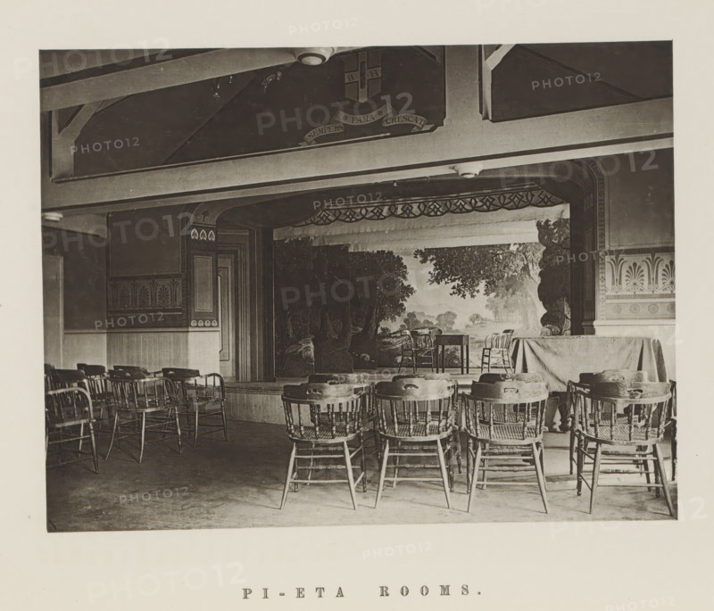 Theatre of the Harvard Pi Eta Society, Pi-Eta rooms, page, photomechanical print, c. 1870 - in or before 1875, paper, collotype, photoprint, height 160 mm x width 210 mm, university building, college, club (students), North America, Massachusetts, Cambridge (MA), Harvard College, A sepia-toned interior photograph showing a small stage with a painted backdrop of trees and a landscape, framed by decorative arches and trim, with two wooden chairs and a small table on the stage and a draped cloth covering a portion of the stage to the right; in front of the stage rows of wooden spindle-back chairs face forward, some grouped around small round tables, and wooden flooring is visible beneath; the ceiling above the stage has exposed beams and a mounted light fixture, and the walls feature patterned friezes and paneling.