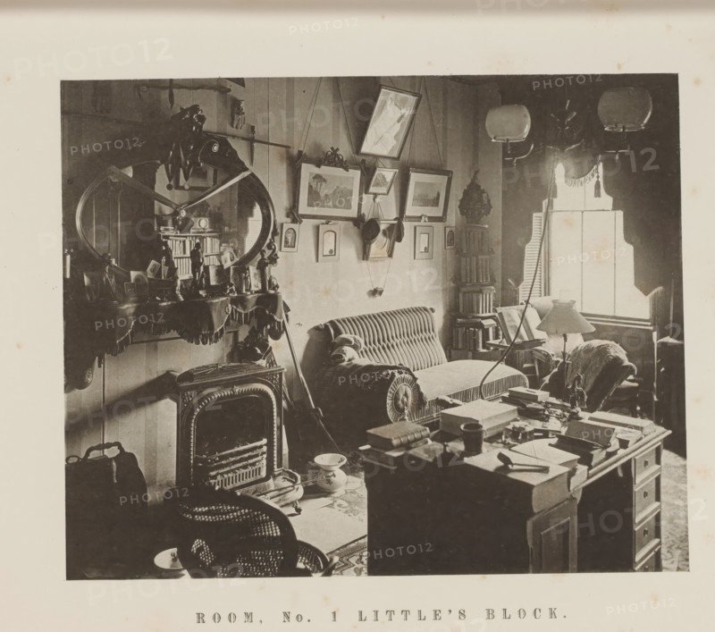Interior of Room One in Little's Block of Harvard University, Room, no. 1 Little's Block, page, photomechanical print, c. 1870 - in or before 1875, paper, collotype, photoprint, height 165 mm x width 195 mm, Print on third unnumbered page after page 48., study, 'studiolo', library, university building, college, North America, Massachusetts, Cambridge (MA), Harvard College, A sepia-toned interior photograph showing a cluttered room with upholstered seating, including a striped sofa with a bolster cushion and an armchair partially covered by a throw, a decorative fireplace with an ornate mirror above it reflecting part of the room, a table in the foreground piled with books and papers, framed pictures hung on the wall at varying angles, a tall narrow bookcase filled with books beside a window with a fringed valance and partially open shutters, three hanging light fixtures with glass shades near the ceiling, various small decorative objects and framed photos on the mantel, a freestanding ...
