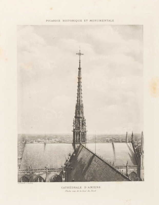 Tower on Notre-Dame of Amiens, page, photomechanical print, inscription: 'Cathédrale d'Amiens, Flèche oue de la tour de Nord', cliché maker: Paul Dujardin, c. 1888 - in or before 1893, Paul Dujardin, photographer, cliché maker, paper, collotype, photoprint, height 210 mm x width 170 mm, parts of church exterior and annexes: tower, Amiens, Notre-Dame d'Amiens, A sepia-toned photograph showing the top portion of a Gothic cathedral with a tall ornate spire rising from the roof, the spire topped by a decorative cross and featuring intricate tracery and sculptural details; the steep sloping roof planes of the cathedral flank the spire, with visible ridge lines, gutters, and rows of pinnacles along the roof edges; the image includes a distant hazy cityscape on the horizon below the roofline and a cloudy sky filling the upper portion of the frame; the overall image appears aged with subtle stains and toning around the paper margins.