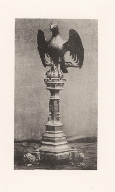 Sculpture of an eagle on a pedestal from the Basilica of Saint Martin in Halle, displayed at an exhibition of religious objects from the Middle Ages and Renaissance in 1864 in Mechelen, page, photomechanical print, number: '-49', number: 'I ...', number: '1', photographer: Joseph Maes, Mechelen, clichémaker: Simonau & Toovey, Antwerp, 1864 - in or before 1866, paper, photolithography, height 238 mm x width 132 mm, predatory birds: eagle, piece of sculpture, reproduction of a piece of sculpture (metals, arts), Period, 1864, A tall sculptural stand topped by a large eagle with outstretched wings perched on a rounded base, the eagle showing detailed feather texture and a forward-facing head, the stand beneath it featuring a multi-tiered column with ornamental moldings and carved panels, and four small lion figures positioned near the base on a flat surface; the scene appears in monochrome with a plain backdrop and visible grain from the photographic print.