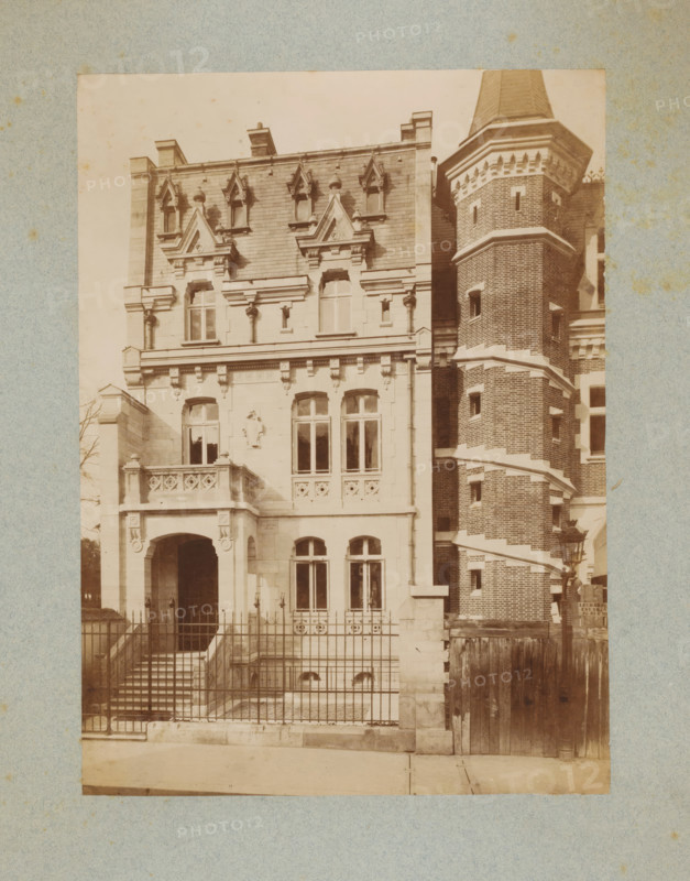 Façade of a building in Paris, photograph, This photo is part of a cover with 35 photos and a title page., number: '1', name: 'De Chiévre. Archte', photographer: Lampué et Cie, Paris, c. 1875 - in or before c. 1880, paper, albumen print, height 226 mm x width 165 mm, façade (of house or building), A sepia-toned photograph of a multi-story building facade showing ornate stonework and a tall brick tower with a conical roof; the left portion of the building features decorative gables, carved stone window surrounds, a small balcony with patterned balustrade, arched entrance framed by columns, and tall windows, while the right side shows a cylindrical brick tower with narrow windows and horizontal stone bands; a metal fence runs across the front with steps leading down to a lower area, and a wooden gate and lamp post are visible near the base of the tower.