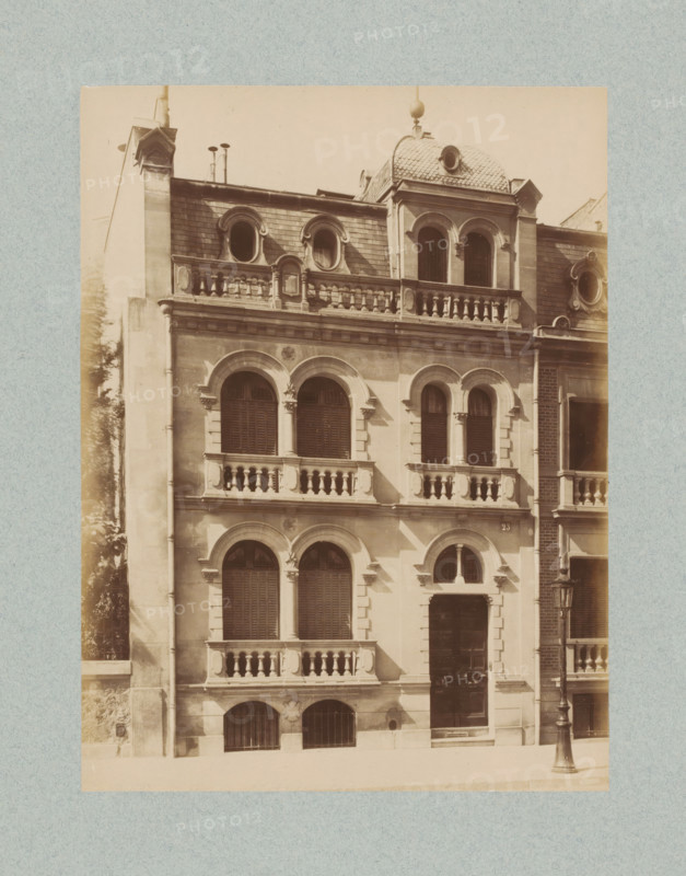 Façade of a building in Paris, photograph, This photo is part of a cover with 35 photos and a title page., name: 'xxx Architecte', number: '12', photographer: Lampué et Cie, Paris, c. 1875 - in or before c. 1880, paper, albumen print, height 221 mm x width 165 mm, urban housing, façade (of house or building), A sepia-toned photograph of a multi-story stone building with arched windows and decorative stone balustrades; the facade shows rows of paired arched windows with shutters and ornamental columns between them, a central entrance recessed beneath an arched surround, and smaller arched basement openings with iron grilles; the uppermost level features rounded dormer windows set into a tiled mansard roof and a small domed roof section with a circular opening, while the roofline includes chimneys and small pipes; a cast-iron street lamp stands on the sidewalk near the building, and the image is bordered by a textured pale background.