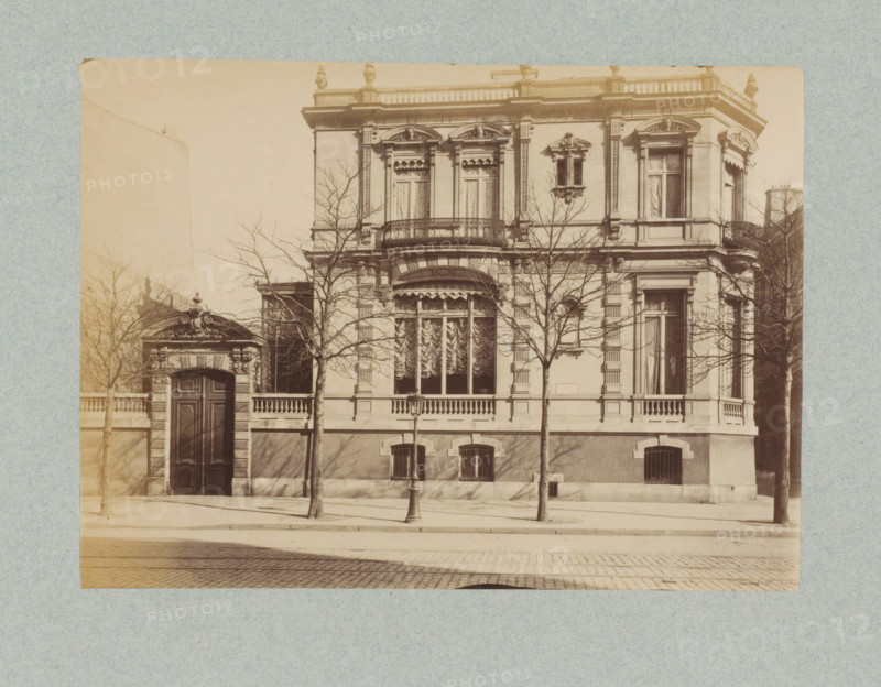 Façade of a villa in Paris, designed by Jules Février, photograph, This photo is part of a cover with 35 photos and a title page., name: 'Février Architecte', number: '14', photographer: Lampué et Cie, Paris, c. 1875 - in or before c. 1880, paper, albumen print, height 165 mm x width 225 mm, detached house in city, façade (of house or building), A sepia-toned photograph of a large ornate building façade with decorative stonework and tall windows partially covered by patterned curtains, a curved balcony with wrought-iron railing above a bay window with swagged drapery, fluted pilasters and ornamental pediments above upper windows, a grand arched wooden doorway set in a small ornate stone gateway at one side, leafless trees planted along the sidewalk whose branches cast shadows on the façade, a lamppost on the pavement in front, small barred basement windows at street level, and a cobblestone street in the foreground.