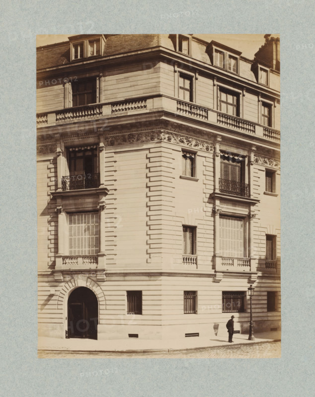 Corner of a building in Paris, photograph, This photo is part of a cover with 35 photos and a title page., name: 'Escalier Architecte', number: '18', photographer: Lampué et Cie, Paris, c. 1875 - in or before c. 1880, paper, albumen print, height 221 mm x width 165 mm, urban housing, A sepia-toned photograph of a multi-story corner building with ornate stonework and decorative cornices, featuring tall windows with shutters and iron balconies, a rounded arched entrance at street level, rusticated horizontal banding on the façade, carved frieze details below the upper balcony, dormer windows in the roof, a streetlamp near the corner, and a man standing on the sidewalk beside the lamp casting a shadow on the wall.