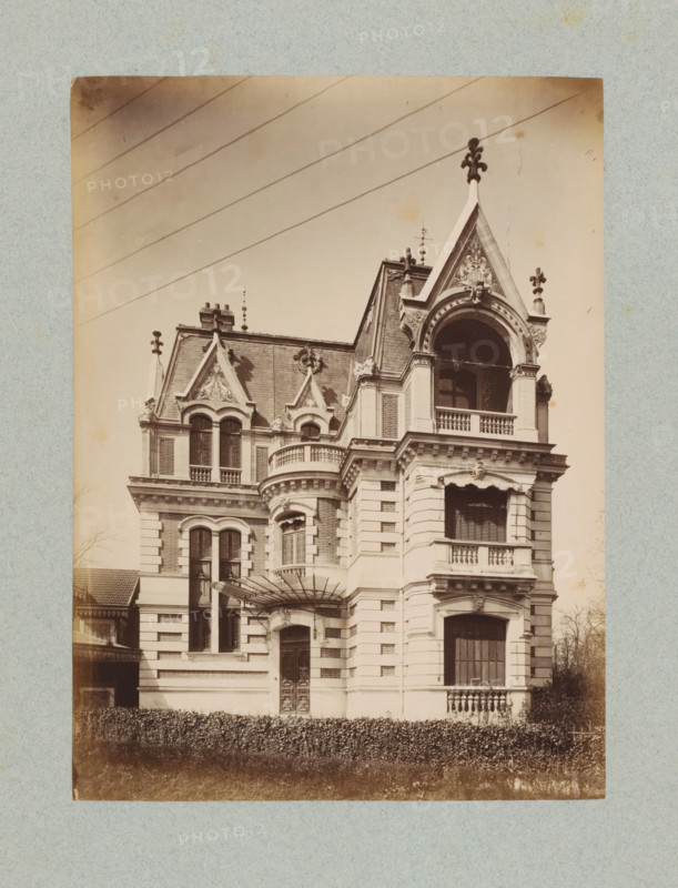 Façade of a villa in Paris, photograph, This photo is part of a cover with 35 photos and a title page., number: '3', name: 'xxx Architecte', photographer: Lampué et Cie, Paris, c. 1875 - in or before c. 1880, paper, albumen print, height 225 mm x width 165 mm, detached house in city, façade (of house or building), A sepia-toned photograph shows the facade of an ornate multi-story building with elaborate stonework and decorative roof gables. The roofline features steep triangular pediments with sculpted reliefs and finials, and multiple dormer windows set into a slate roof. A large arched opening with a balustraded balcony occupies an upper corner, framed by carved moldings and columns. Below, stacked balconies with balustrades and shuttered windows are set into a rusticated exterior with alternating smooth and textured stone bands. Tall arched windows with decorative tracery appear on one side, and a carved doorway with an awning and glass canopy is visible at ground level. A neatly tr...