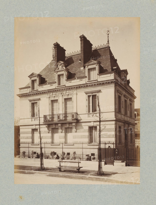 Façade of a villa in Paris, designed by Albert-Ernest Carrier-Belleuse, photograph, This photo is part of a cover with 35 photos and a title page., number: '4', name: 'Carrier Architecte', photographer: Lampué et Cie, Paris, c. 1875 - in or before c. 1880, paper, albumen print, height 220 mm x width 165 mm, detached house in city, façade (of house or building), A sepia-toned photograph of a multi-story European-style house with a steep mansard roof, ornate dormer windows, decorative stonework around the windows and cornice, and tall brick chimneys rising from the roofline. The facade shows shuttered windows, a small wrought-iron balcony above the entrance, and sculpted details beneath the roof edge. In front of the house a metal fence runs along the sidewalk, with two bare young trees protected by circular grates and metal guards. A wooden bench sits on the pavement near the trees and a gated pedestrian entrance is visible to the side. The image has a textured border and shows even lig...