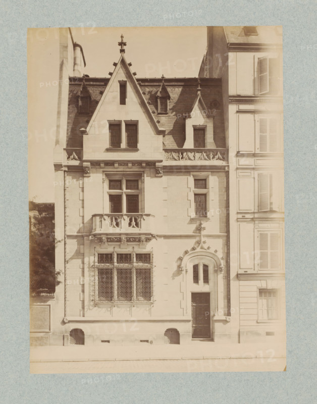 Façade of a residential house in Paris, designed by Henri Deverin, photograph, This photo is part of a cover with 35 photos and a title page., name: 'Deverin Architecte', number: '8', photographer: Lampué et Cie, Paris, c. 1875 - in or before c. 1880, paper, albumen print, height 224 mm x width 165 mm, urban housing, façade (of house or building), A sepia-toned photograph showing the façade of a multi-story stone building with steep gabled roofs and decorative roof finials, narrow dormer windows set into dark slate roofing, ornate carved stone balustrades and window surrounds, and tall rectangular windows with leaded or lattice glazing and external metal grilles. A rounded arched doorway with sculpted embellishments sits beside a tall dark wooden door, and a small balcony with carved supports projects beneath a pair of upper windows. The right edge of the image shows the adjacent building with shuttered windows and paneled surfaces, while the left edge shows a partial view of a chimney...