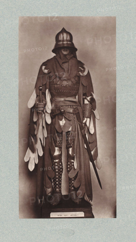 14th-century armor from the army of John II of France, from the collection of the Musée d'Artillerie in Paris, photomechanical print, This print is part of a cover with 36 prints, a title page and a list of images., check stamp: 'PHOTOGLYPTIE, LEMERCIER ET Cie, PARIS', Paris, cliché maker: Lemercier & Cie., c. 1877 - in or before 1882, Lemercier & Cie., photographer, cliché maker, paper, Woodburytype, Woodburytypie, height 270 mm x width 131 mm, armor, A full-length sepia-toned photograph of a person standing on a small platform wearing an ornate costume that includes a rounded helmet with a flared brim obscuring the eyes, a layered cloak with scalloped and teardrop-shaped fabric pieces hanging from the shoulders and sleeves, and a high wrapped collar covering the neck and lower face. The outfit features patterned leggings with decorative studs or spots, metal knee guards, and studded footwear. The person holds a long sword in one hand and a staff or mace-like object in the other, with...