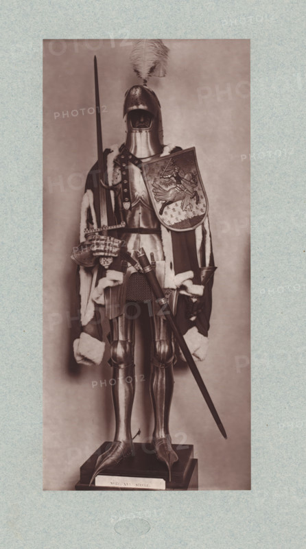 15th-century armor with sword and shield from the army of Charles VII of France, from the collection of the Musée d'Artillerie in Paris, photomechanical print, This print is part of a cover with 36 prints, a title page and a list of images., check stamp: 'PHOTOGLYPTIE, LEMERCIER ET Cie, PARIS', Paris, cliché maker: Lemercier & Cie., c. 1877 - in or before 1882, Lemercier & Cie., photographer, cliché maker, paper, Woodburytype, Woodburytypie, height 275 mm x width 127 mm, hacking and thrusting weapons: sword, armour, protective weapons: shield, A full suit of medieval-style armor displayed on a pedestal, shown front-facing with a closed helmet topped by a large feather plume, a breastplate and articulated arm and leg armor, and gauntleted hands; a long spear is held upright in one hand while a decorated shield with an embossed scene rests against the torso; a fur-trimmed cloak drapes over the shoulders and down the sides, and a sword and scabbard hang at the waist; the armor stands on a...